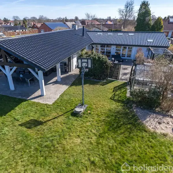 En have med en græsplæne indeholder et fodboldmål og en basketballkurv. Ved siden af står en carport med overdækning og et hus i baggrunden.