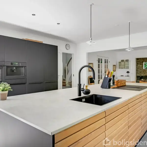 Køkkenø med lys bordplade og integreret vask; står i et moderne køkkenrum med mørk skabssektion, planter, og knive på blok. Vogner og hængende lamper skaber en minimalistisk atmosfære.