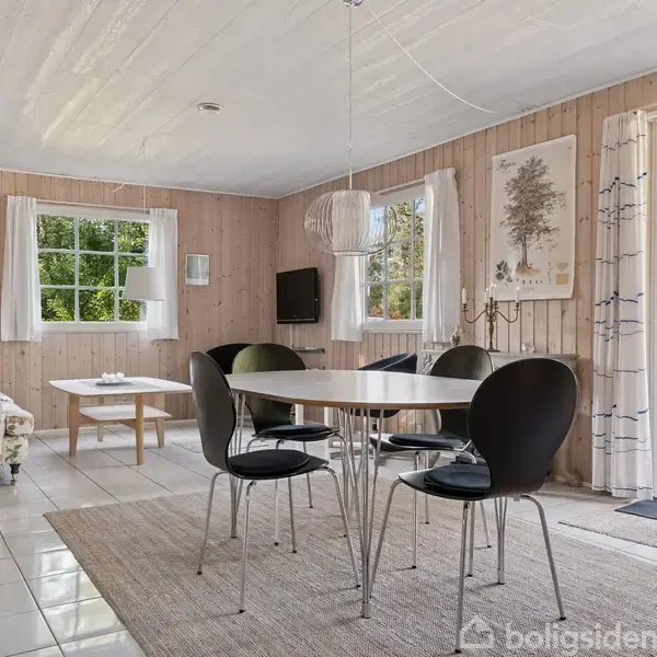 Et spisebord med fire sorte stole står på et gråt tæppe i en stue med lyse trævægge og hvide fliser. Store vinduer og en åben dør giver udsigt til naturen.
