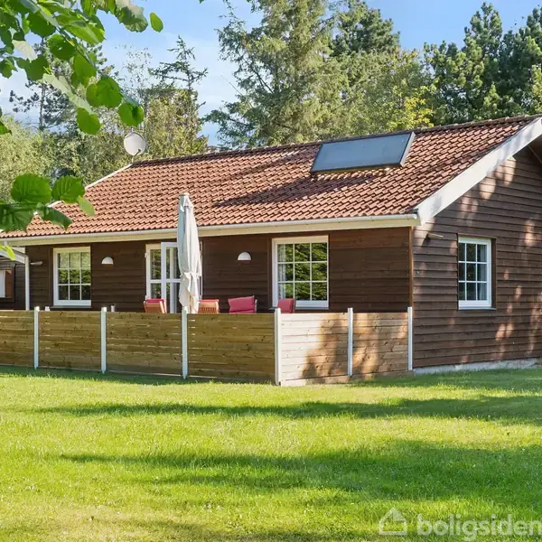 Et træhus med rødt tegltag står stille i en grøn have. Der er en terrasse med en parasol, omgivet af træer.