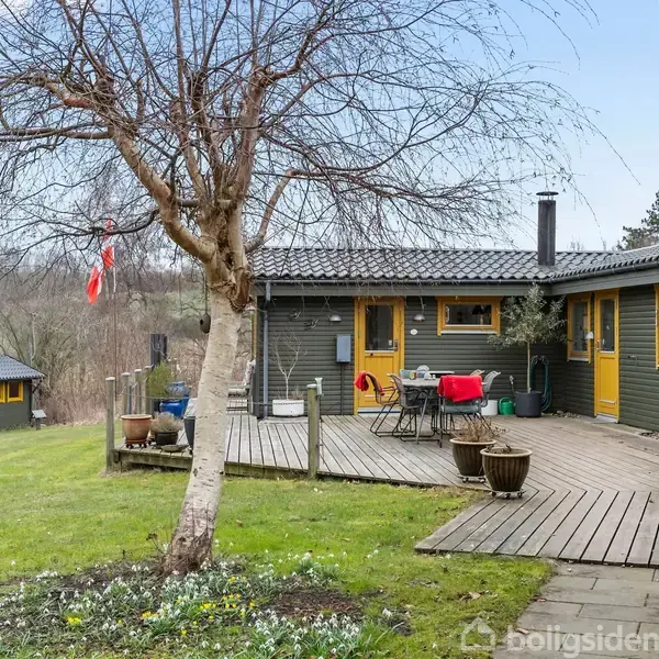 Et træ står i en have foran et træhus med en terrasse. Havemøbler og planter er placeret på terrassen. Et mindre skur ses i baggrunden, omgivet af græsplæne og marker.