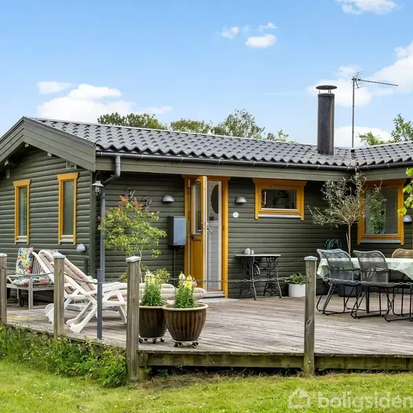 Træhus står på træterrasse med havemøbler og planter i krukker, omgivet af grøn græsplæne og træer under en klar blå himmel.