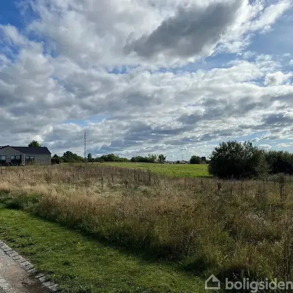En lav bygning står stille ved en vej med en græsmark og buske omkring. Ovenover er himlen fyldt med skyer.