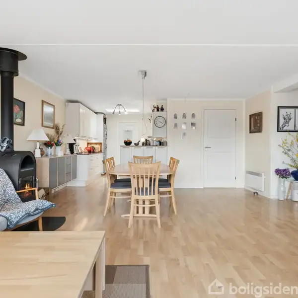 Et lyst opholdsrum med spisebord og stole i midten. En brændeovn står til venstre ved en grå sofa. Køkkenet ses bagerst med hvidt interiør. Væggene er dekoreret med kunst.