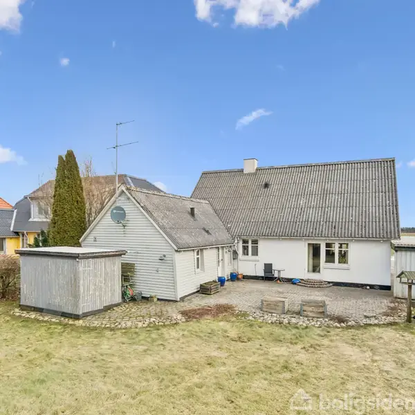 Et hvidt hus med gråt tag står i en have med græsplæne og buske. Der er et lille skur ved siden af. Himlen er blå med få skyer.