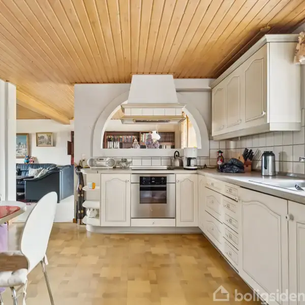 Et hvidt køkken med indbyggede skabe og moderne apparater. Køkkenet åbner mod en stue med synligt træloft og spisebord. Vindue til højre giver naturligt lys og udsigt.