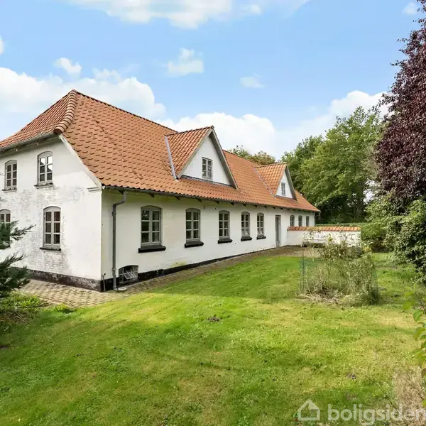 Hvidt hus med rødt tegltag står stille på en grøn græsplæne. Der er træer og buske i baggrunden og til siderne under en blå himmel med skyer.