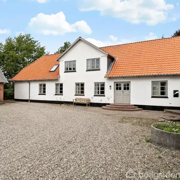 Et hvidt stuehus med rødt tegltag står på en gårdsplads belagt med grus. Til venstre ses en ældre lade i røde mursten. Omkring er grøn beplantning.