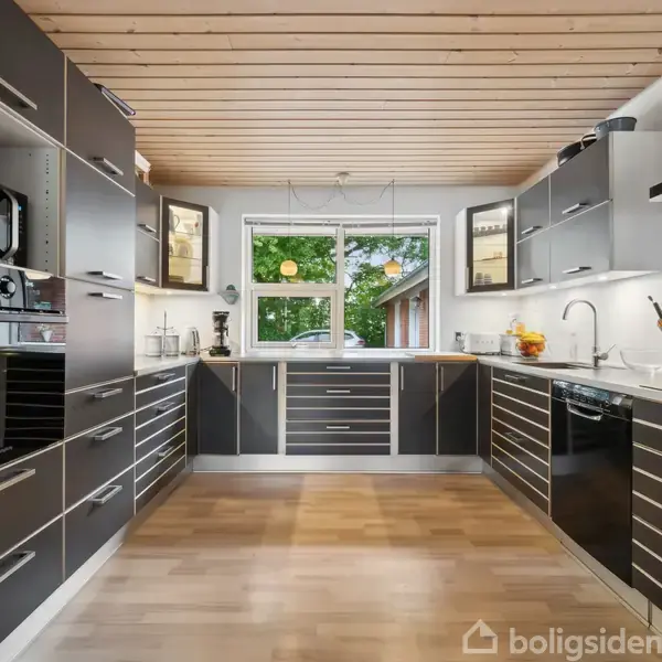 Moderne køkken med mørke skabe, hvor opvaskemaskinen er i fuld gang. Det ligger i et rum med trægulv og træloft, med udsigt til en have gennem et stort vindue.