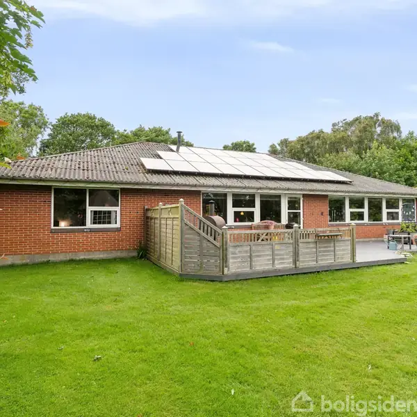 En lav, rød murstensvilla med solpaneler på taget har en træterrasse foran. Villaen er omkranset af en grøn græsplæne og træer.