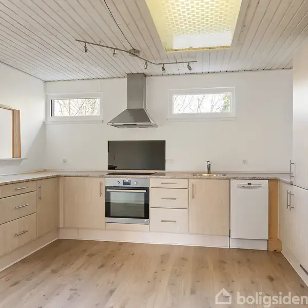 Et moderne køkken med lyse træskabe, indbygget ovn og emhætte. Rummet har lyse trægulve og et vindue, der giver naturligt lys. En opvaskemaskine er integreret i skabslinjen.