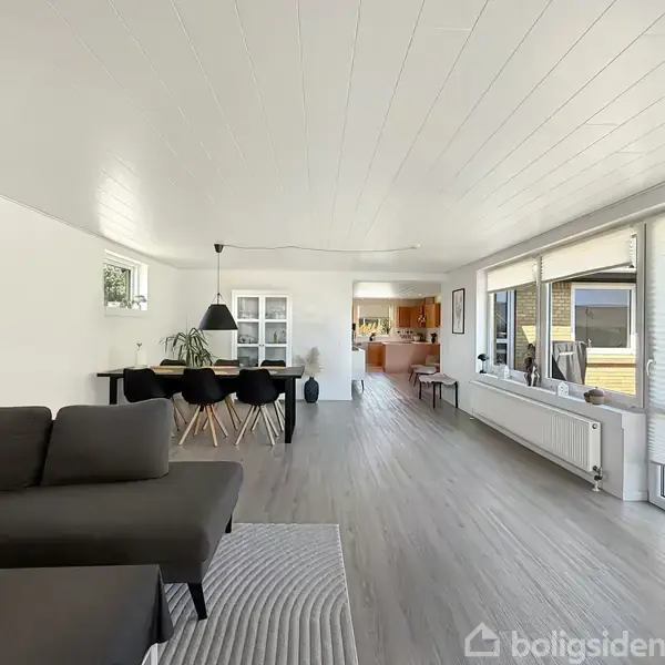 Gråt sofaarrangement står stille i en moderne, lys stue med trægulve. Til venstre er et spisebord med sorte stole. Store vinduer giver udsigt til en terrasse.