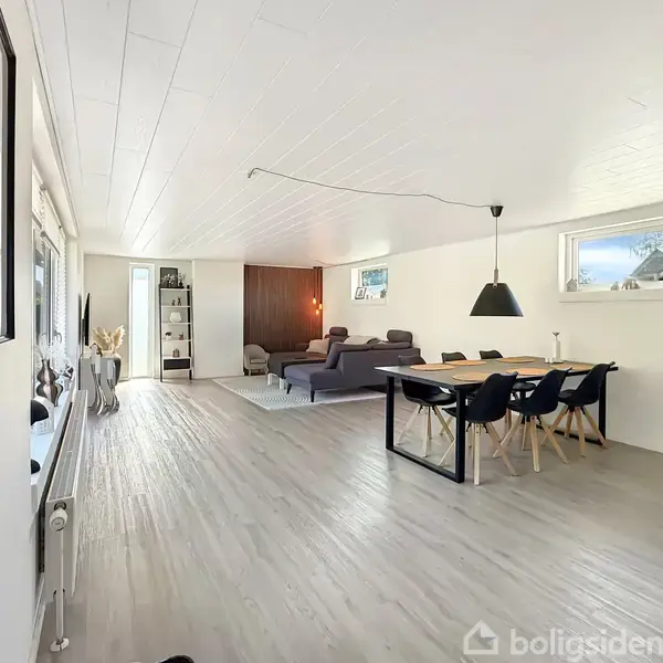 Stue, minimalistisk indrettet, med en grå sofa og et spisebord med sorte stole, placeret i et lyst rum med trægulv og hvidmalede vægge.