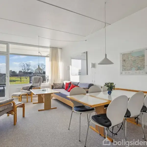 Stue møbleret med sofa, stole og spisebord. Rummets store vindue viser udsigt til grønt landskab. Et kort hænger på væggen. Møblerne er lyse med neutralt interiør.