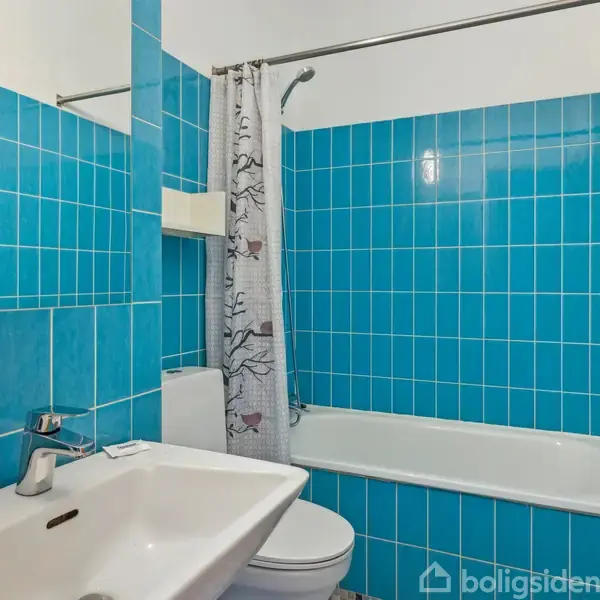 Et badeværelse med blå fliser har en håndvask med sæbedispenser, et spejl, toilet, og badekar med bruseforhæng. En dør står åben, og der hænger et blåt håndklæde.