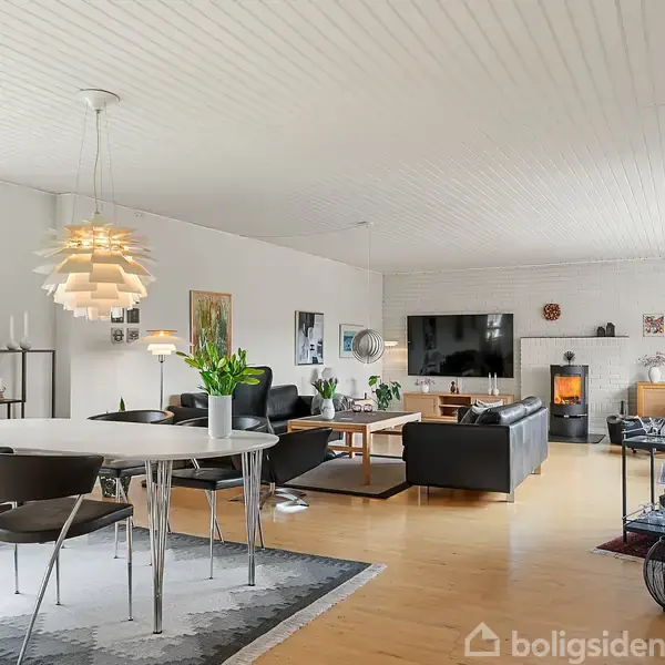Spisebord med stole står i en moderne stue, hvor der er et ophængt lysarmatur. I baggrunden er en pejs, sofaer og et tv på væggen, omgivet af planter og dekorationer.