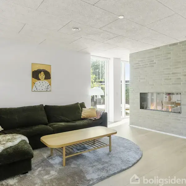 Sort sofa med puder står ved siden af en væg med moderne pejs. Lys kommer ind fra store vinduer, og et billede hænger på væggen. Stuebordet står på tæppe.