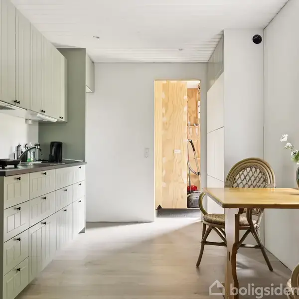Et køkken har lyse træskabe og en sort bordplade. Et træbord med kurvestole står ved vinduet med udsigt til haven. En vase med blomster pynter bordet.