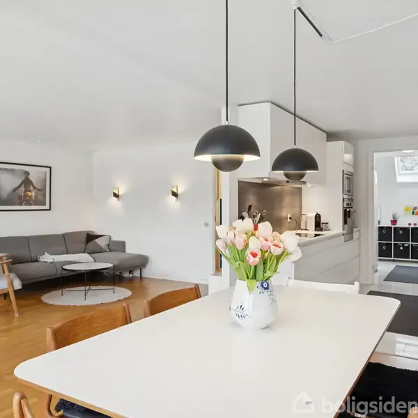 Et lyst køkken-alrum med et bord med blomster i en vase, omgivet af stole. Ved siden af er en sofagruppe med et kaffebord og kunst på væggen.