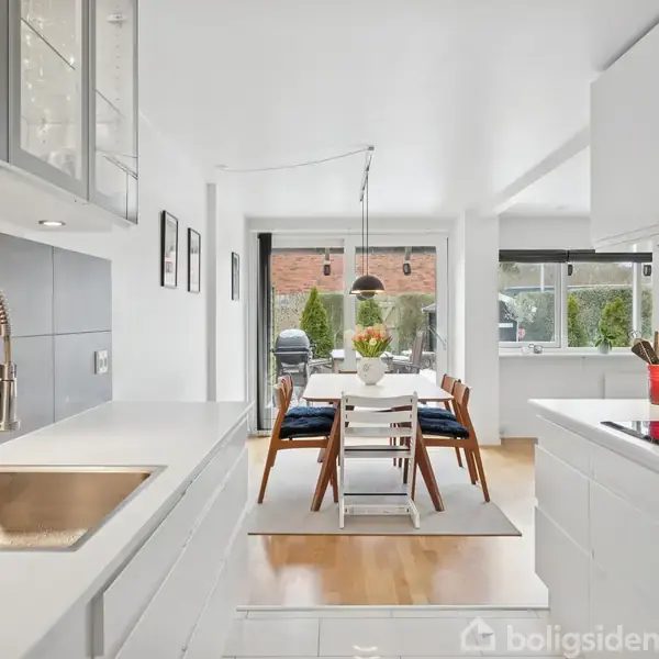 Køkken med hvidt interiør og stålvask. Spisebord med fire blåstole og en vase i midten. Store vinduer i baggrunden slipper lys ind. 여성걸컬렉션.