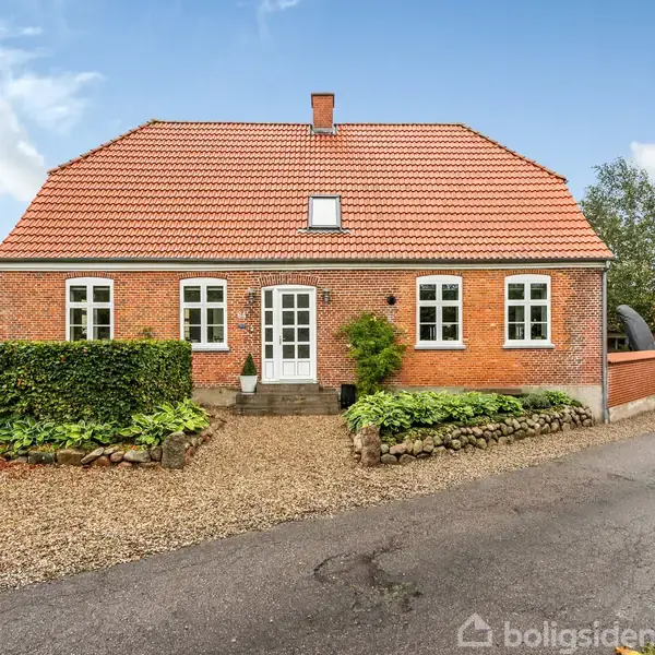 Rødt murstenshus med rødt tegltag og hvide vinduer. Foran huset er der en grusvej og stenbed, omkranset af lav buskbeplantning. Huset ligger i et grønt, landligt område.