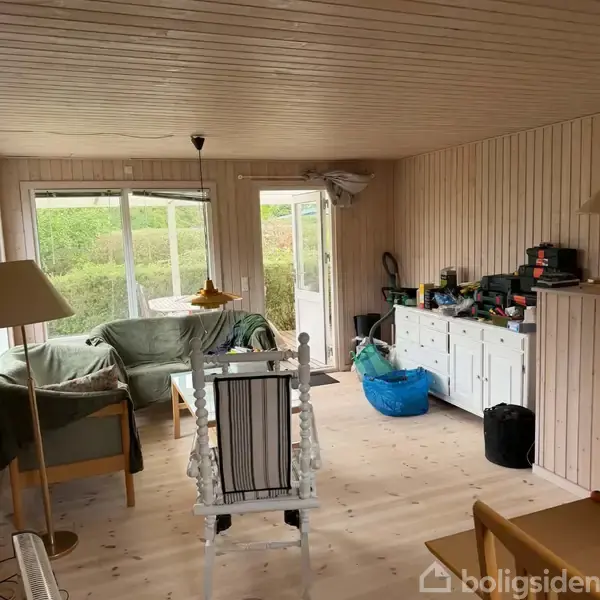 Stue med trævægge og loft. En sofa og bord til venstre, dør mod terrasse i midten, og skænk med ting til højre. Lyse omgivelser.