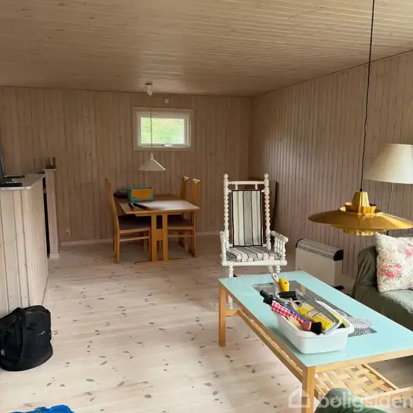 Stue med lyse træpaneler. Et spisebord står til venstre med stole omkring. Foran, en gyngestol ved et sofabord med værktøj og tæpper. En grøn sofa med en standerlampe ved siden af.