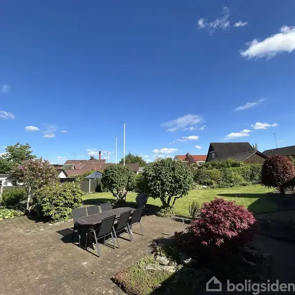 Terrassemøbler står på en solrig terrasse med udsigt over en velplejet have omkranset af buske og træer. I baggrunden ses flere hustage mod en klar blå himmel.