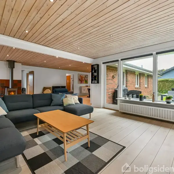 Sofaen er anbragt i en stue med et træloft. Den står ved et sofabord på et ternet tæppe. Store vinduer giver udsigt til en terrasse med liggestole.