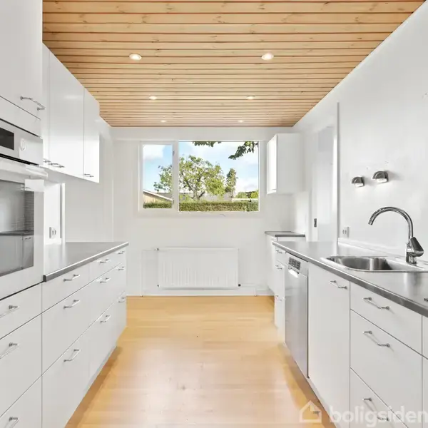 Hvidt køkken med moderne ovn og skabsmoduler på begge sider, står stille. Træloft med spotlys, vindue bagved viser grøn udsigt. Ingen tekst i billedet.