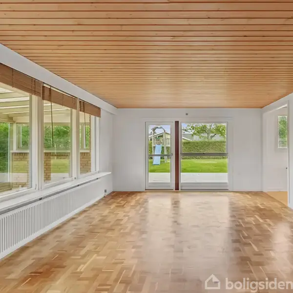 En stor, tom stue med trægulv og loft i lyst træ. Store vinduer og glasdøre åbner ud til en grøn have i baggrunden.