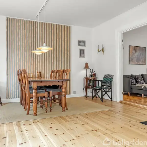 Et spisebord med stole står i en spisestue. Loftslampe lyser over bordet. Rummet har lyse trægulve og en væg med lodrette trælister. I baggrunden ses en sofa i stuehjørnet.