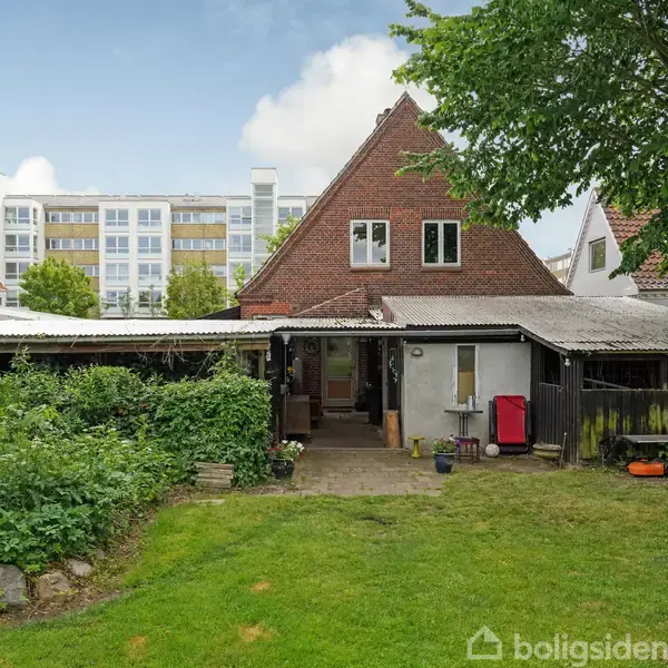 Rødt murstenshus med stejl gavl står i en have med plæne og buske. Foran huset er der en overdækket terrasse med havemøbler. I baggrunden ses en moderne bygning.