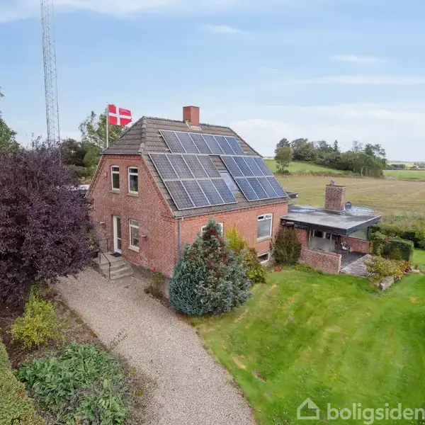 Et rødt murstenshus med solpaneler på taget står ved siden af en have. En dansk flagstang er placeret ved siden af huset. Omgivelserne er landlige og grønne.