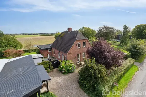 Rødt murstenshus med sort tag står omgivet af træer og buske; en grusvej fører til ejendommen, som ligger i et landligt område med marker i baggrunden under en blå himmel.