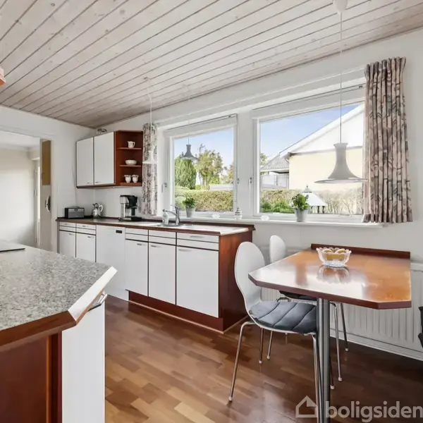 Køkken med hvide skabe og træbordplade, står roligt under et lyst loft. Bord med stole placeres ved store vinduer med udsigt til have.