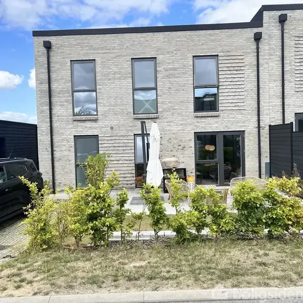 To-etagers hus med grå mursten, åbne døre og parasol i gårdhaven. En sort bil parkeret til venstre og hække foran. Klar blå himmel med få skyer ses i baggrunden.