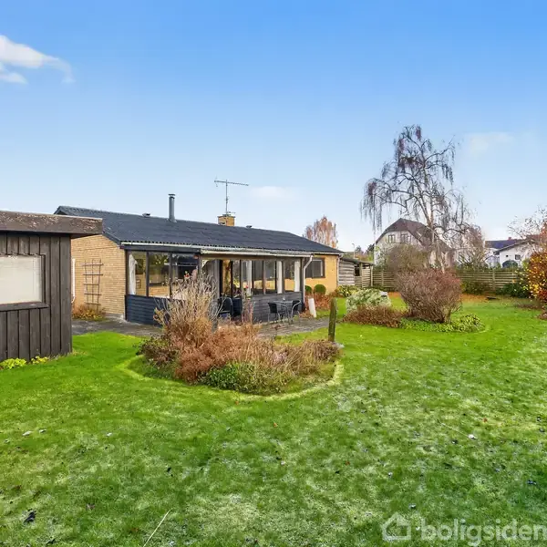 En enkeltstående, sort træhus og et murstenshus med glastilbygning står i en velplejet have med grønne græsplæner og buske under en klar blå himmel.