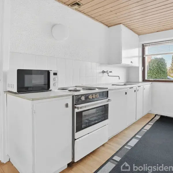 Køkken med hvidt kabinet, komfur og mikrobølgeovn på bord. Sollys kommer ind gennem vinduet. Træloftet og lyse vægge giver et skandinavisk udtryk. Tæppe på gulvet.