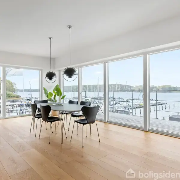 Et spisebord med seks stole står i et rummeligt rum med store vinduer. Udsigt over en marina med både udenfor. To hængelamper hænger over bordet. En plante pynter midten.