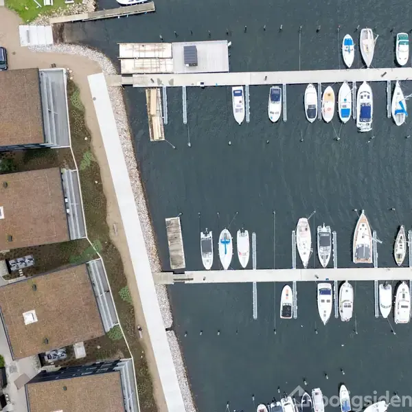 Både ligger fortøjet ved en lang bådebro i en marina. Til venstre ses flere huse med flade tage omgivet af træer og veje.