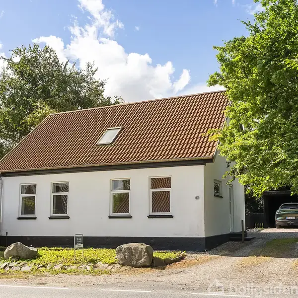 Hus med hvid facade og rødt tag står ved vejen. En grusindkørsel fører til en carport med en bil. Omgivet af træer og buske.