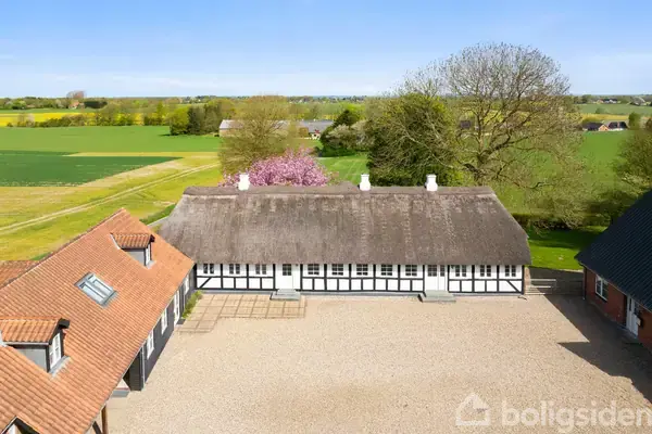 Bindingsværksbygning med stråtag står stille i en gårdsplads omgivet af grus. Omgivet af marker og træer, under en klar blå himmel. Blomstrende træer ses i baggrunden.