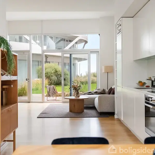 Reol med planter står ved siden af en lys stue med sofabord og sofa. Store vinduer vender ud mod en terrasse med grønne planter udenfor. Køkkenet er til højre.