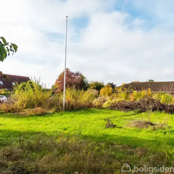 Flagstang står stille i en villahave omgivet af grønne buske og træer. Baggrunden viser to huse med brune tage under en blå, let overskyet himmel.