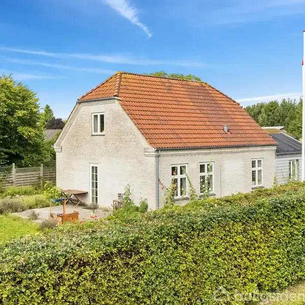 Et hvidt hus med rødt tegltag står i en grøn have med buske. Et bord og stole står udenfor. Et flag flagrer på høj flagstang, omgivet af træer og hæk.