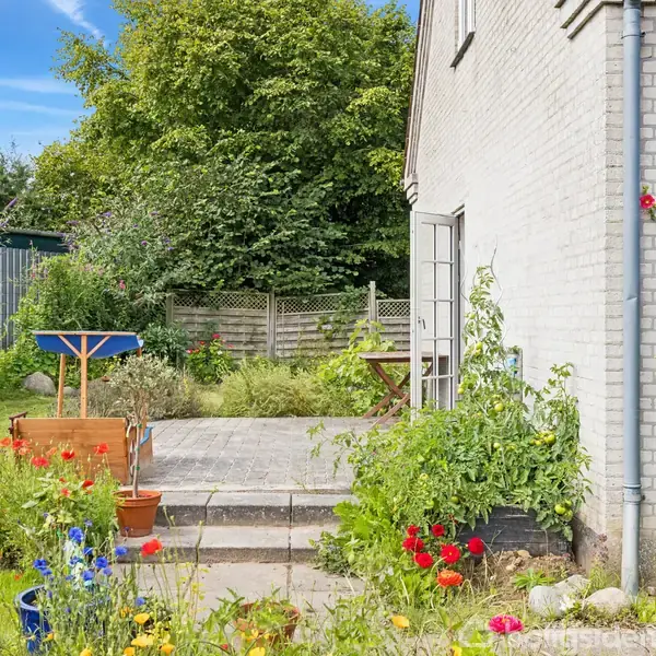 Hus med lys murstensfacade og åben dør ud til terrasse, omkranset af farverige blomsterbede og grøn beplantning. Blå himmel og frodig have skaber en sommerlig atmosfære.