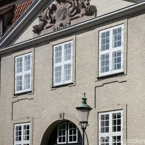 Facade af en ældre bygning med tre store hvide vinduer. En gadelampe står foran bygningen, der er lavet af lyse mursten. Tallet "9" er placeret over en dør.