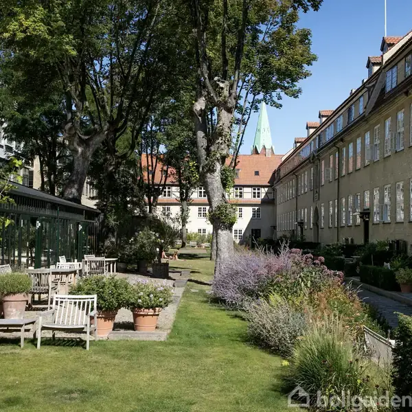 En gårdhave med træbænke og blomsterkrukker placeret blandt grøn græsplæne. Omkranset af bygninger med røde tegltage og store vinduer. Store træer kaster skygger over området.