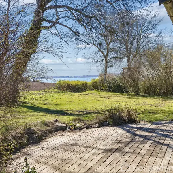 Træterrasse grænser op til en græsplæne med træer i baggrunden. I baggrunden ses havudsigt og blå himmel.